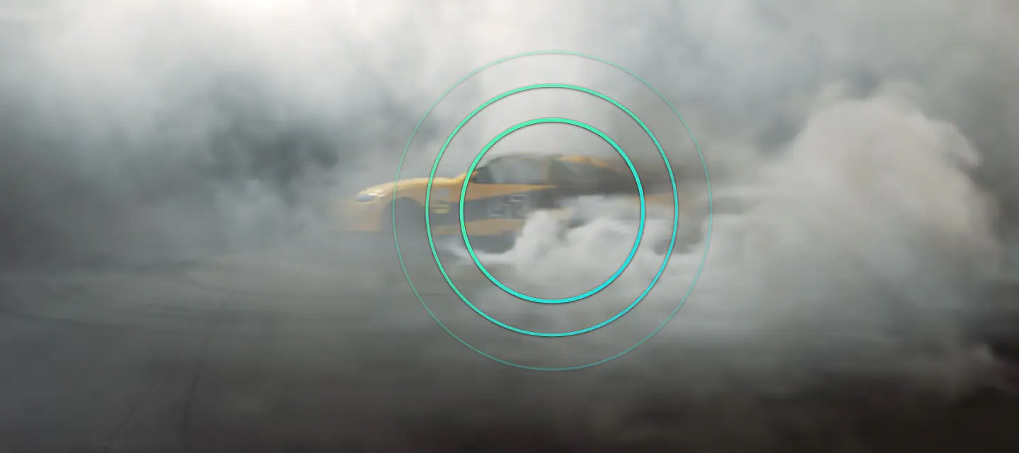 A wide shot view of a race track shows a race car speed into view, spinning its tires as it drifts in a counter-clockwise motion. The smoke from the spinning tires rises from the race track. From the moment the race car enters the screen to the moment it exits, an animated ripple graphic can be seen superimposed on top of the race car to illustrate Neo QLED's Object Tracking Sound Pro feature which allows the race car's sound to be heard from its location on the screen.